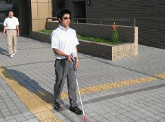 写真。歩行訓練の様子。2人の人物が訓練中。遮光眼鏡を付けた利用者が、順光に向かいながら視覚障害者用誘導ブロックに沿って、白杖を用いて歩いている。その後ろから、指導員が、歩行の様子を確認しつつ歩いている。